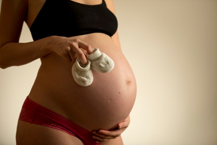 Foto mulher grávida segurando um pequeno par de botinhas de tricô sobre a barriga nua enquanto segura a barriga com a outra mão, vista de perto das mãos com espaço de cópia