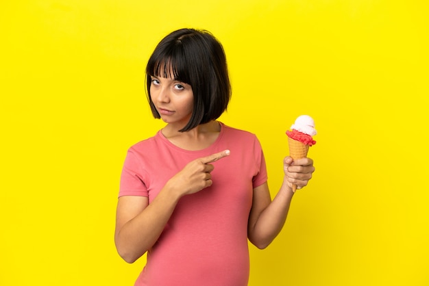 Mulher grávida segurando um sorvete de corneta isolado em um fundo amarelo apontando para o lado para apresentar um produto