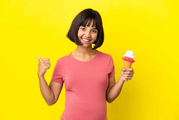 Mulher grávida segurando um sorvete de corneta isolado em um fundo amarelo apontando para o lado para apresentar um produto