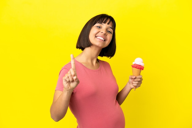 Mulher grávida segurando um sorvete de corneta isolado em um fundo amarelo, mostrando e levantando um dedo