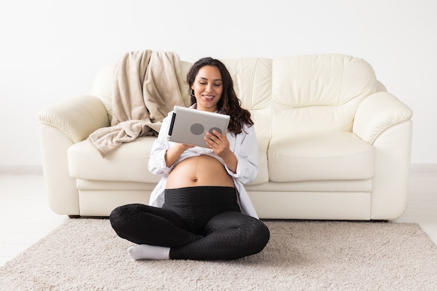 Mulher grávida segurando um tablet sentada em um tapete perto de um sofá na sala de estar de casa