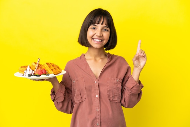 Mulher grávida segurando waffles isolados na superfície amarela apontando o dedo para o lado e apresentando um produto