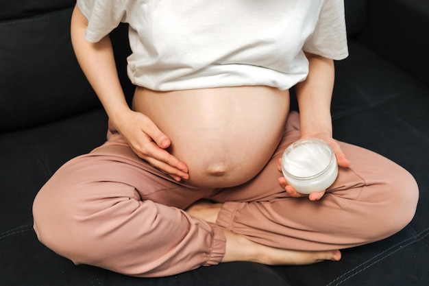 Mulher grávida sentada na cama acariciando aplicar creme na barriga grande em casa
