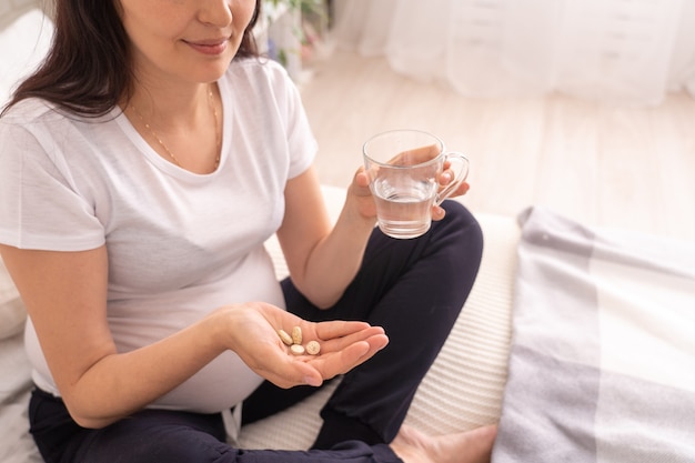 Mulher grávida sentada na cama e segurando um copo de água potável e vitaminas