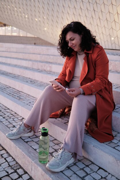 Mulher grávida sentada nas escadas na cidade relaxando segurando o telefone gravando histórias da viagem