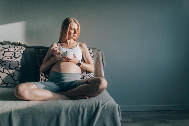 Mulher grávida sentada no sofá e segurando um prato com ricota e mirtilos