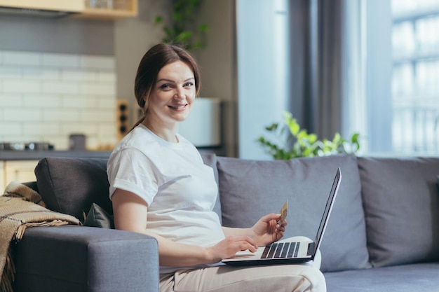 Mulher grávida sentada no sofá em casa usando laptop para fazer compras na loja online olhando para a câmera e sorrindo segurando o cartão de crédito do banco