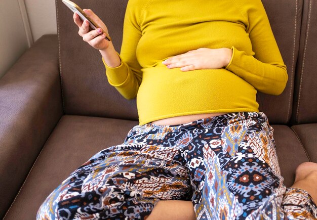 Mulher grávida sentada no sofá usando o celular mulher grávida assistindo conteúdo de mídia em um telefone sentada em um sofá na sala de estar em casa conteúdo autêntico da vida real