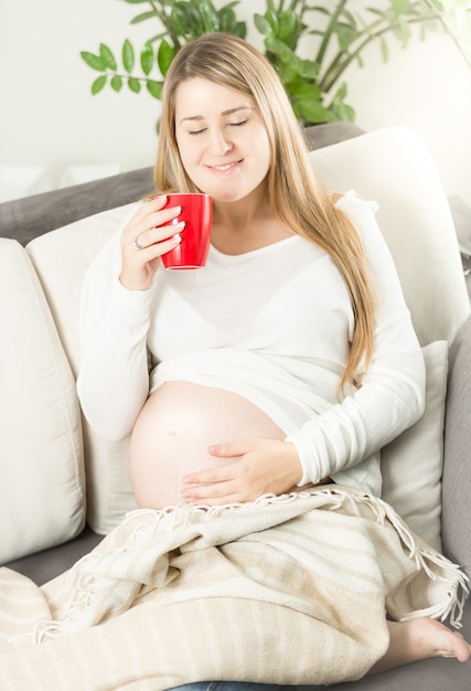 Mulher grávida sorridente deitada no sofá e bebendo chá