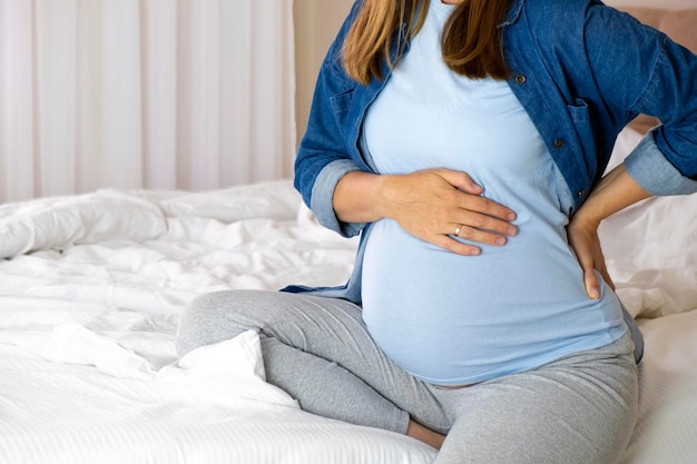 Mulher grávida toca sua barriga e mantém uma mão nas costas sentada na cama em seu quarto Dor nas costas dor nas costas dores de parto saúde gravidez problemas de saúde