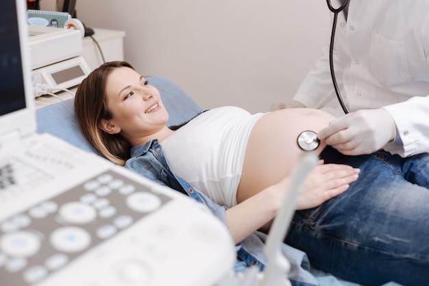 Mulher grávida tranquila, sorridente, desfrutando de uma consulta no hospital e fazendo um exame de barriga enquanto obstetra usa o estetoscópio para verificar os batimentos cardíacos do feto