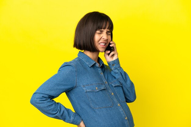 Mulher grávida usando celular isolado em fundo amarelo, sofrendo de dor nas costas por ter feito esforço