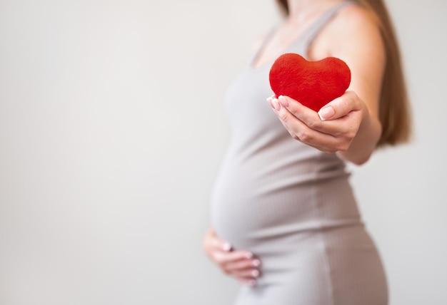 Mulher grávida usando vestido apertado segurando coração vermelho macio em fundo cinza Conceito de saúde amor gravidez Ideia de problemas de cardiologia ou doençaCopiar espaço Vista lateral corpo desfocado