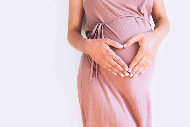 Mulher grávida vestida de mãos dadas na barriga sobre um fundo branco. Maternidade na gravidez