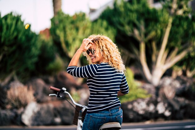 Mulher hippie com cabelos cacheados e óculos escuros, sentada na bicicleta, olhando por cima do ombro e admirando algo interessante. mulher feliz andando de bicicleta e admirando algo distante