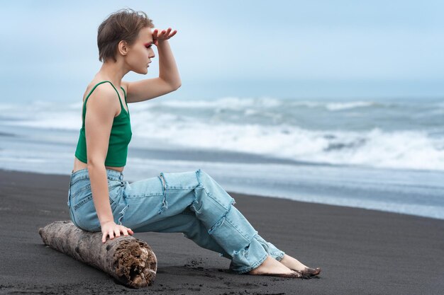 Mulher hippie sentada em um tronco na praia de areia preta do oceano levantou a mão na testa olhando a vista