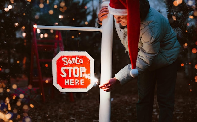 Mulher hippie usando um chapéu de papai noel à noite com um diamante acende uma placa de pare com o texto papai noel pare aqui.