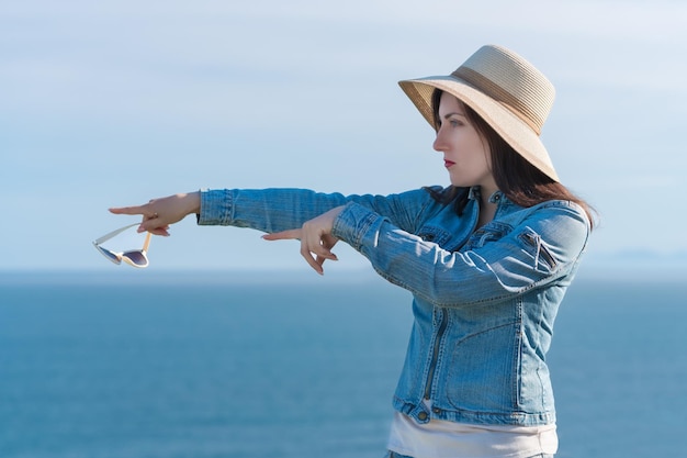 Mulher hipster com chapéu de palha e jaqueta jeans mostra com as duas mãos a direção para o lado