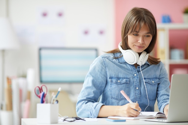 Mulher hipster de conteúdo em camisa jeans sentada à mesa e fazendo anotações no organizador enquanto trabalha wi