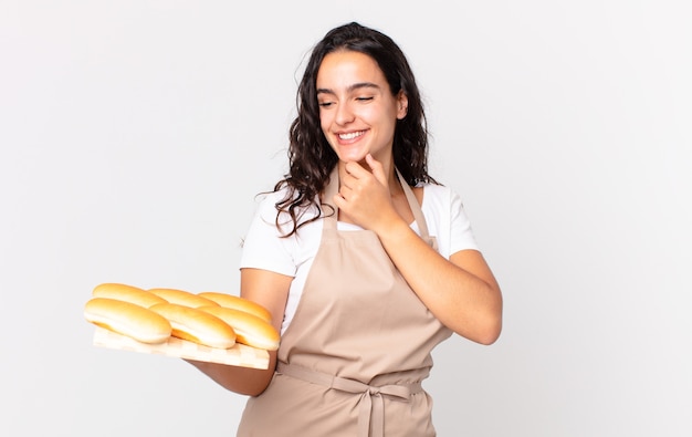 Foto mulher hispânica bonita chef sorrindo com uma expressão feliz e confiante com a mão no queixo e segurando um troy de pães