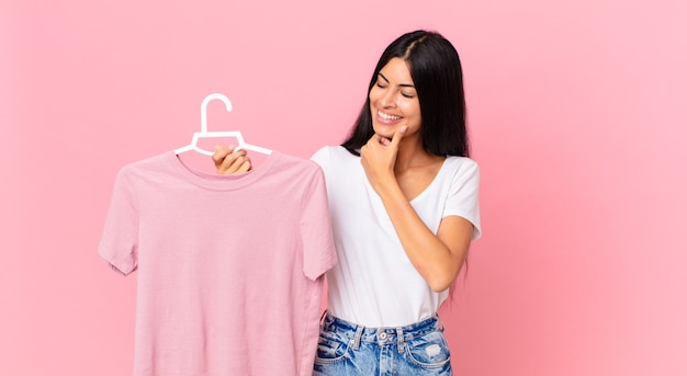 Mulher hispânica bonita sorrindo com uma expressão feliz e confiante com a mão no queixo e segurando um pano escolhido