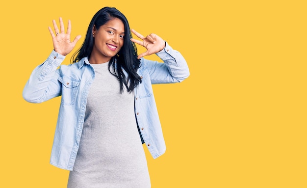 Mulher hispânica com cabelo comprido vestindo jaqueta jeans casual mostrando e apontando para cima com os dedos número oito enquanto sorria confiante e feliz.