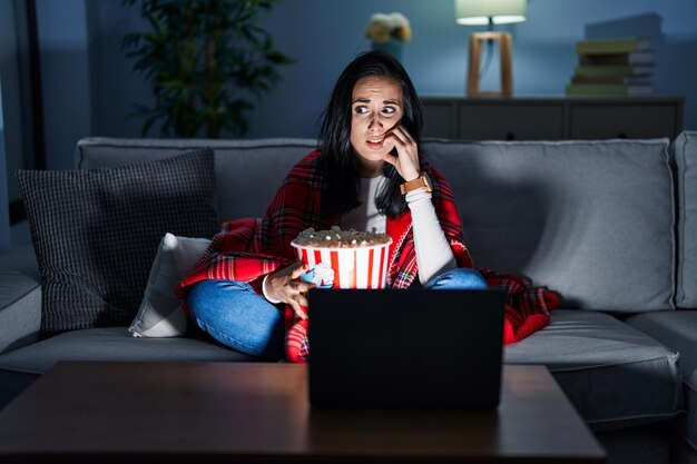 Foto mulher hispânica comendo pipoca assistindo a um filme no sofá parecendo estressada e nervosa com as mãos na boca, roendo unhas, problema de ansiedade
