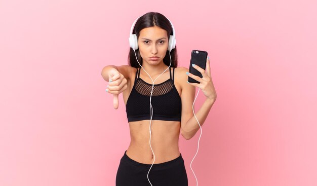 Mulher hispânica de fitness se sentindo mal, mostrando os polegares para baixo com fones de ouvido e um telefone