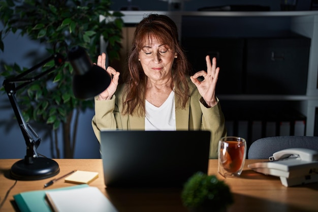Mulher hispânica de meia-idade trabalhando usando computador portátil à noite relaxar e sorrir com os olhos fechados fazendo gesto de meditação com os dedos conceito de ioga