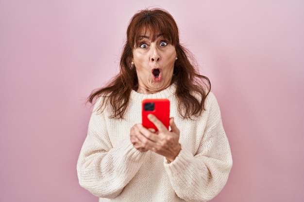 Mulher hispânica de meia-idade usando mensagem de digitação de smartphone com medo e chocada com surpresa e expressão espantada, medo e rosto animado.