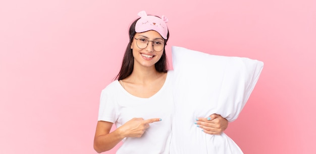 Mulher hispânica de pijama sorrindo alegremente, feliz, apontando para o lado e segurando um travesseiro