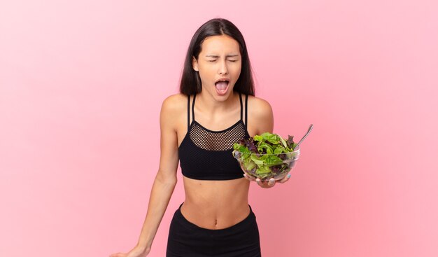 Mulher hispânica em boa forma gritando agressivamente, parecendo muito zangada e segurando uma salada