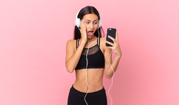 Mulher hispânica em fitness se sentindo feliz, animada e surpresa com fones de ouvido e um telefone