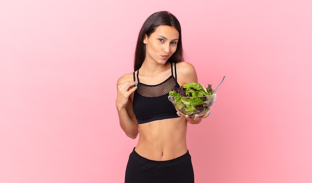 Mulher hispânica em forma de fitness parecendo arrogante, bem-sucedida, positiva e orgulhosa, segurando uma salada