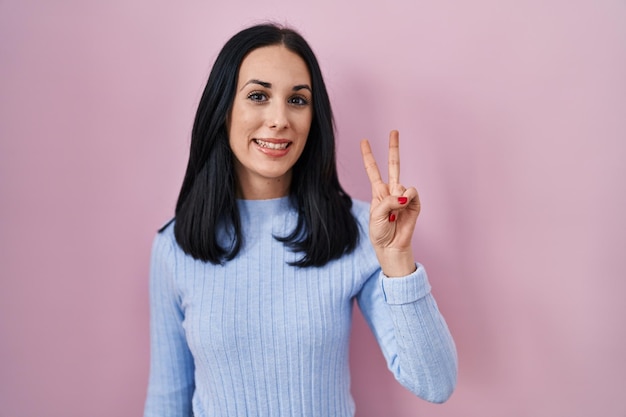 Mulher hispânica em pé sobre fundo rosa mostrando e apontando para cima com os dedos número dois enquanto sorria confiante e feliz.