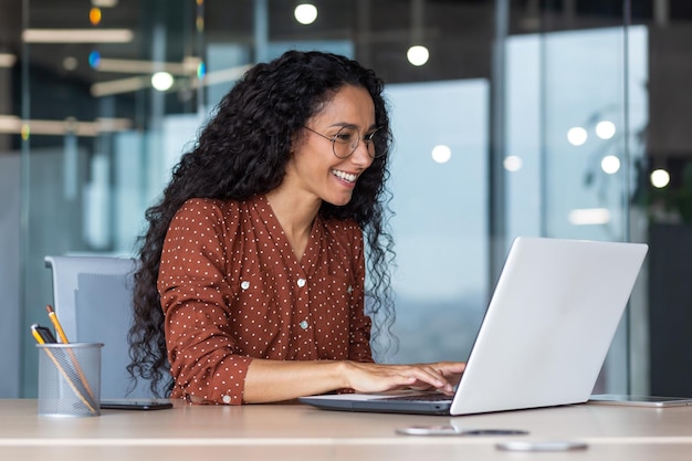 Mulher hispânica jovem e feliz trabalhando em escritório moderno usando mulher de negócios de laptop sorrindo e