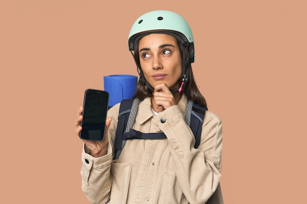 Mulher hispânica mostra telefone com capacete olhando para o lado com expressão duvidosa e cética