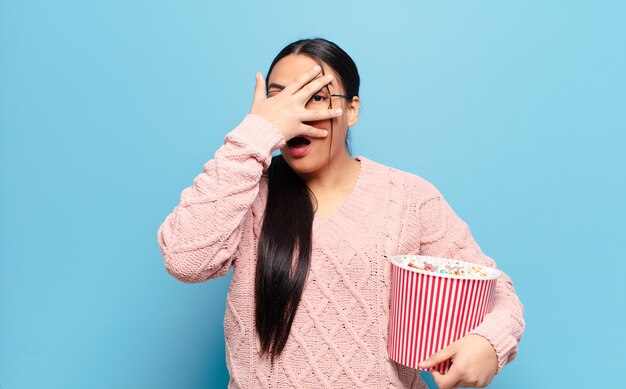 Mulher hispânica parecendo chocada, assustada ou apavorada, cobrindo o rosto com a mão e espiando por entre os dedos