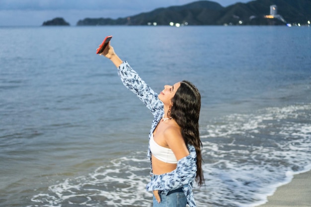 Mulher hispânica rindo enquanto toma selfie com celular na praia