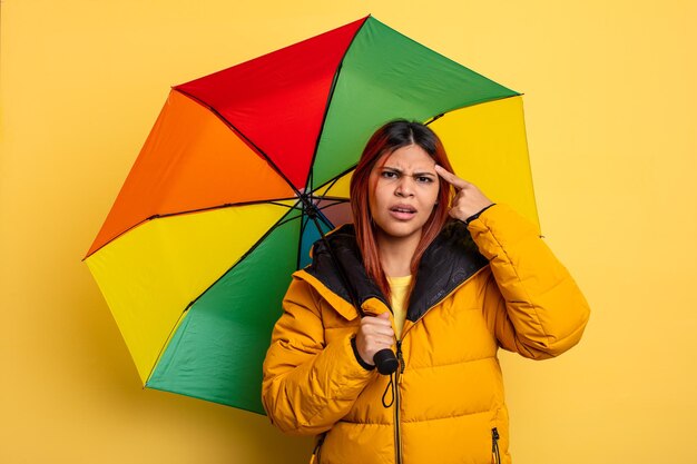 Mulher hispânica se sentindo confusa e confusa, mostrando que você é louco. conceito de chuva e guarda-chuva