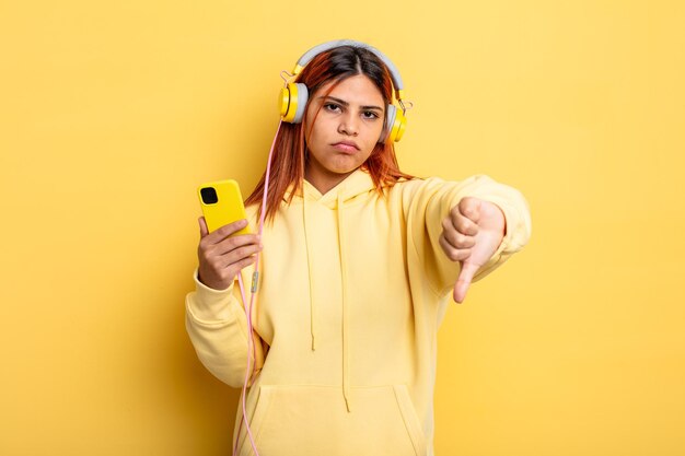 Mulher hispânica se sentindo zangada, mostrando os polegares para baixo. fones de ouvido e conceito de smartphone