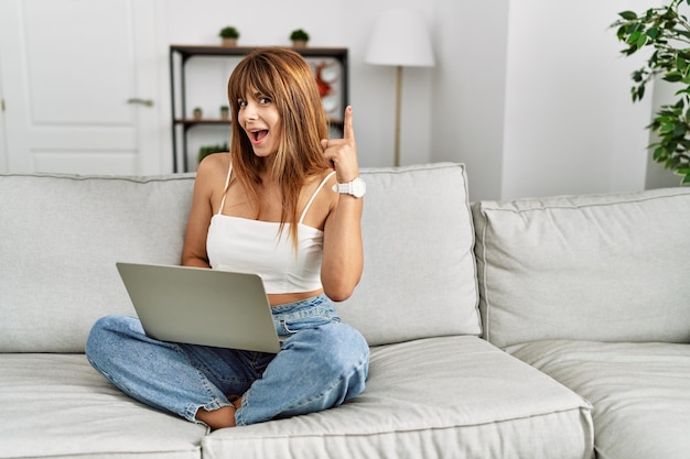 Mulher hispânica sentada no sofá em casa usando laptop apontando o dedo para cima com uma ideia bem-sucedida e feliz número um