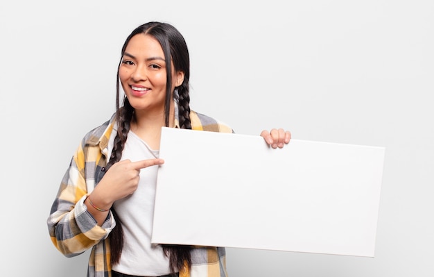 Mulher hispânica sorrindo alegremente, sentindo-se feliz e apontando para o lado e para cima, mostrando o objeto no espaço da cópia