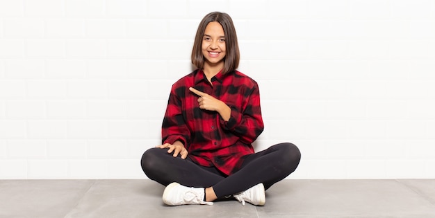 Mulher hispânica sorrindo alegremente, sentindo-se feliz e apontando para o lado e para cima, mostrando o objeto no espaço da cópia