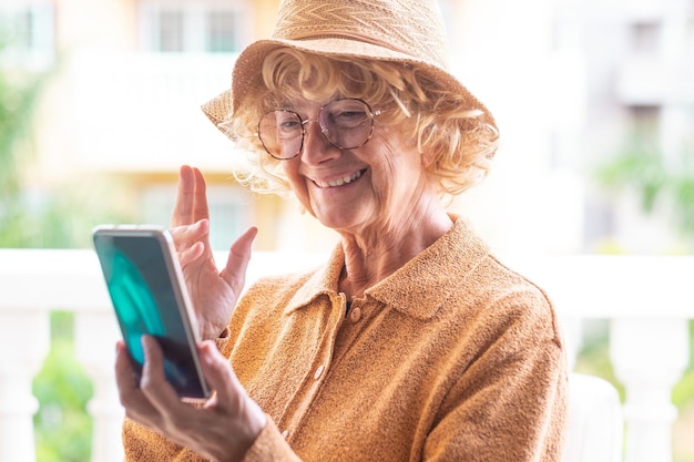 Foto mulher idosa alegre de vestido amarelo com chapéu e óculos ao ar livre em videochamada com telefone celular loira encaracolada mais velha desfrutando de tecnologia e social