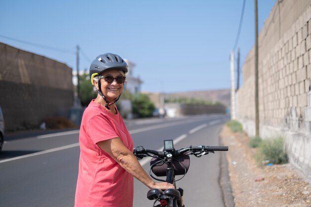 Mulher idosa alegre usando capacete de proteção correndo em sua bicicleta elétrica na estrada