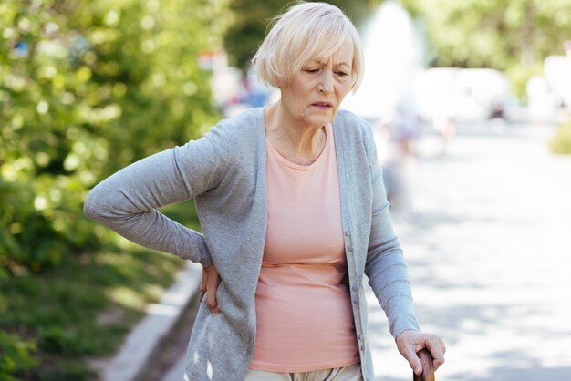 Foto mulher idosa aposentada deprimida tocando suas costas e apoiando-se na bengala enquanto sofre de dores nas costas ao ar livre