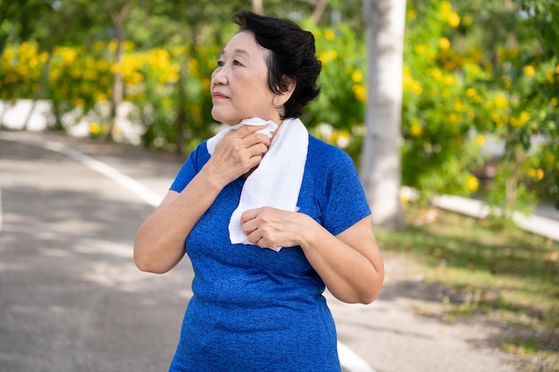 Mulher idosa asiática cansada usando toalha para enxugar o suor enquanto caminhava ou fazia jogging no parque