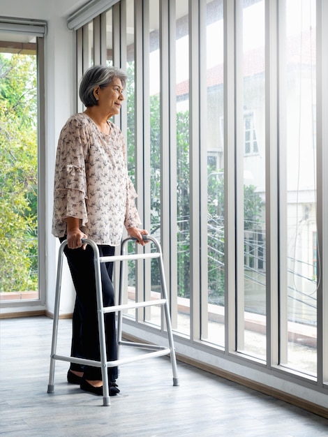 Mulher idosa asiática de cabelo branco em pé com armação de caminhada de comprimento total olhando pela janela de vidro estilo vertical Paciente idosa usando armação de caminhada Conceitos de cuidados médicos de saúde fortes