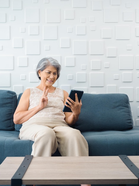 Mulher idosa asiática feliz senta-se no sofá fazendo o sinal da vitória com a mão na tela do tablet digital na mão estilo vertical Mulher idosa se encontra com a família por videochamada em casa Pessoas mais velhas com tecnologia